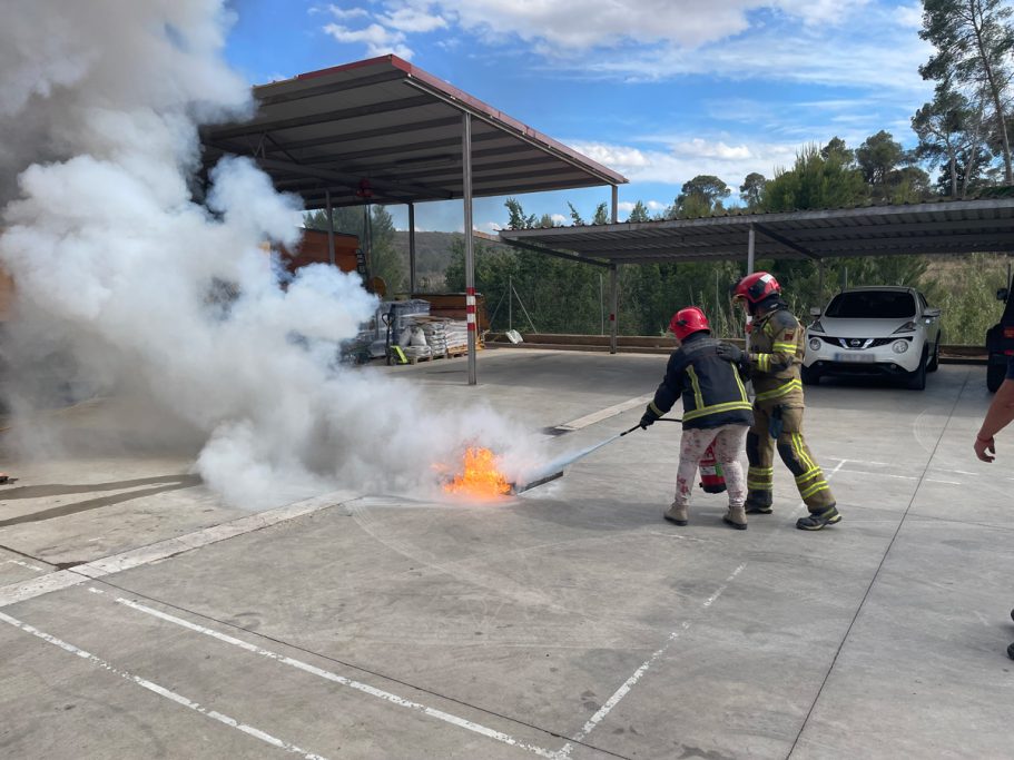 Mujer apagando un fuego con extintor
en un área de entrenamiento, tras ella Bombero instructor.