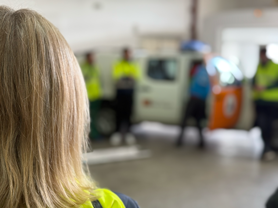 Mujer de espaldas observa a un grupo de instructores y participantes cerca de un vehículo de emergencias.