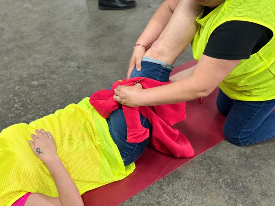 Mujer realizando un vendaje en la pierna de otra persona que está en el suelo, Lo realiza con una prenda de ropa.